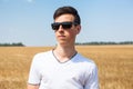 Teenage Caucasian boy portrait with black sunglasses, man stands on wheat field at sunny day, white t-shirt