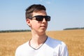 Teenage Caucasian boy portrait wearing black sunglasses, man standing on a wheat field at sunny day, white t-shirt