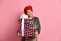 Teenage female in gray pajamas, red hat. She holding colorful gift boxes, showing tongue, posing on pink background. Close up