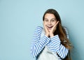 Teenage female in jeans overall, striped sweatshirt. She smiling, showing thumbs up, winking, posing on blue background. Close up