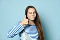 Teenage female in jeans overall, striped sweatshirt. She smiling, showing thumbs up, winking, posing on blue background. Close up