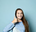 Teenage female in jeans overall, striped sweatshirt. She smiling, showing thumbs up, winking, posing on blue background. Close up