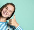 Teenage female in jeans overall, striped sweatshirt. She smiling, showing thumbs up, winking, posing on blue background. Close up