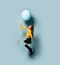 Teenage female in yellow sweatshirt, black skirt, knee-highs, boots. She holding balloon, posing sideways on blue background.