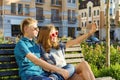 Teenage friends girl and boy sitting on bench in city, talking, looking in the phone, doing selfie photo Royalty Free Stock Photo