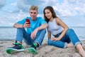 Teenage friends guy and girl sitting together on beach, talking looking at smartphone screen Royalty Free Stock Photo