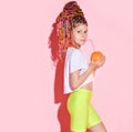 Teenage girl with african pigtails is holding orange and drinking it through tubule, posing sideways on pink background. Close up