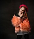 Teenage girl with african pigtails, in red hat, orange faux fur coat. Propping her chin by forefinger, posing on dark background