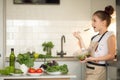 A teenage girl in an apron holds a bowl of self-made salad and tries it. On the kitchen table are green vegetables, red tomatoes a