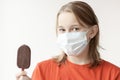 A teenage girl with blond hair, a medical mask on her face looks at the camera, holding a chocolate ice cream in her hand, Royalty Free Stock Photo