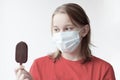 A teenage girl with blond hair, a medical mask on her face looks at a chocolate ice cream that she holds in her hand, selective Royalty Free Stock Photo