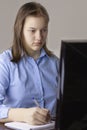 A teenage girl in a blue shirt looks at the computer monitor and makes notes in a notebook that lies on the table next to the