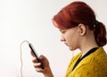 A teenage girl with bright red hair, wearing small headphones, wearing a yellow knitted jacket, in profile, looking at the phone.