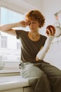 teenage girl with broken arm in cast in kitchen sitting on countertop eating apple