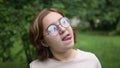 A teenage girl with glasses makes funny faces at the camera in the park. Foolishness and fun. Frames in 4K format