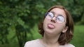 A teenage girl with glasses makes funny faces at the camera in the park. Foolishness and fun. Frames in 4K format