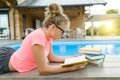 Teenage girl in glasses reads a book, background swimming pool, lawn near the house. School, education, knowledge, adolescents