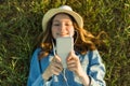 Teenage girl in hat with headphones lies on the green grass and looks into the phone. Focus on smartphone, top view. Royalty Free Stock Photo