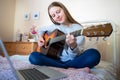 Teenage Girl Learning To Play Acoustic Guitar With Online Lesson On Laptop Computer