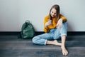 Teenage girl looking on cellphone and smiles sitting on wooden floor. Beautiful girl in yellow sweater and blue jeans holding mobi