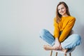 Teenage girl looks at camera .sitting on white modern chair with legs up. Beautiful girl in yellow sweater and blue jeans in home Royalty Free Stock Photo