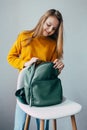 Teenage girl looks inside leather rucksacks that lies on a white modern chair. Beautiful girl in yellow sweater and blue jeans sta