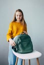 Teenage girl looks on leather rucksacks and to fasten a backpack that lies on a white modern chair. Beautiful girl in yellow sweat