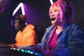 Teenage girl with pink braid using headset competing with her friend in online gaming tournament. Purple lighting.