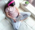 Teenage girl in pink headphones using tablet pc sitting on white couch at home. Top view. Distance learning, quarantine,