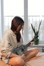 Teenage girl playing ukulele sitting on a bed in the bedroom against the window