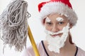 A teenage girl in a Santa hat and with a beard, mustache and eyebrows made of shaving foam, face close-up, holding a MOP.