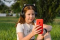 A teenage girl sits in a park on the grass with a smartphone in her hands. 12 year old girl listening to music in headphones Royalty Free Stock Photo