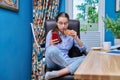 Teenage girl sitting on armchair near computer, looking in smartphone, eating snack Royalty Free Stock Photo