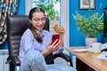 Teenage girl sitting on armchair near computer, looking in smartphone, eating snack Royalty Free Stock Photo