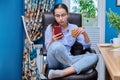 Teenage girl sitting on armchair near computer, looking in smartphone, eating snack Royalty Free Stock Photo