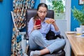 Teenage girl sitting on armchair near computer, looking in smartphone, eating snack Royalty Free Stock Photo