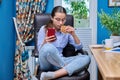 Teenage girl sitting on armchair near computer, looking in smartphone, eating snack Royalty Free Stock Photo