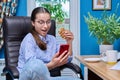 Teenage girl sitting on armchair near computer, looking in smartphone, eating snack Royalty Free Stock Photo