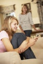 Teenage Girl Sitting On Sofa At Home Texting On Mobile Phone With Mother In Background Royalty Free Stock Photo