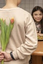 A teenage girl is sitting at a table, a boy hides a bouquet of flowers behind his back Royalty Free Stock Photo