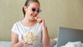 Teenage girl is watching movie on laptop and eating popcorn while sitting in bed. Young happy woman watching movie or TV Royalty Free Stock Photo