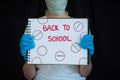 Teenage girl wearing face mask and surgical gloves, holding up notebook that says Back to School