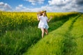 Teenage girl in white dress and Ukrainian wreath runs through yellow fields and green meadows, against cloudy sky Royalty Free Stock Photo