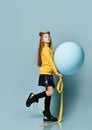 Teenage girl in yellow sweatshirt, black skirt, knee-highs, boots. She holding balloon, posing sideways on blue background.