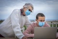 Teenage grandson with grandmother wearing mask because of coronavirus sitting outdoors at a wooden table looking at the same