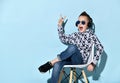 Teenage kid in headphones, sunglasses, blue jeans, hoodie, sneakers. Showing victory sign, sitting on white chair. Blue background Royalty Free Stock Photo