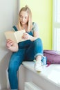 Teenage schoolgirl reading book and sitting on window sill with soda