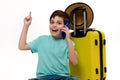 Teenage traveler boy in blue t-shirt, with yellow suitcase, talks on mobile phone, points forefinger at copy ad space