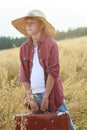 Teenage traveler in farm oat field holding old-fashioned suitcase and looking to horizon