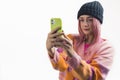 Teenage white girl with pink dyed hair in colorful sweater and beanie taking selfie with her smartphone. Isolated white